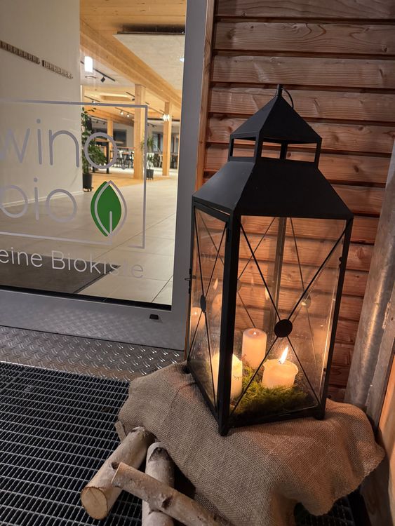 Laterne mit brennenden Kerzen vor einem Holzhaus und Glastür mit Bio-Siegel im Innenraum