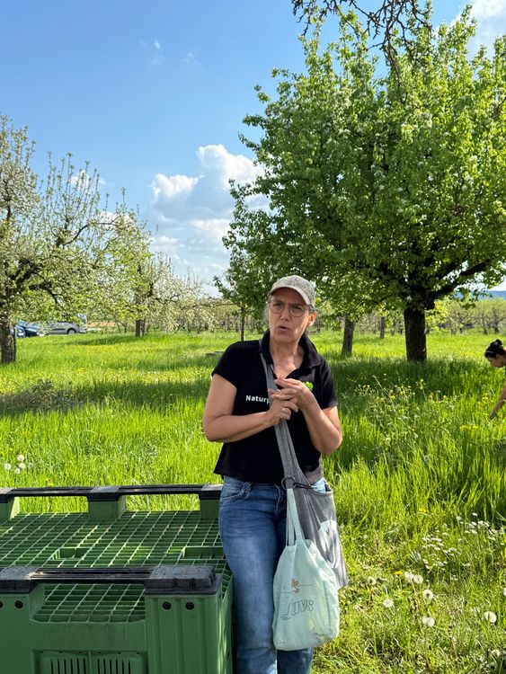 Frau mit Cap und Brille steht auf grüner Wiese neben grünem Behälter, hinter ihr Bäume und blauer Himmel