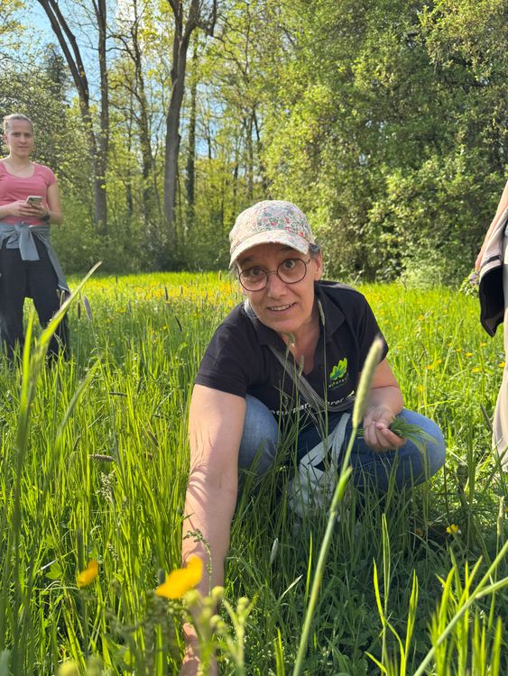 Frau mit Blumenmütze und Brille kniet auf einer grünen Wiese, andere Menschen im Hintergrund