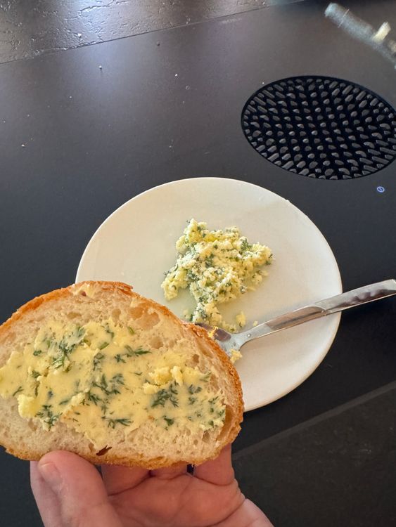 Hand hält Brotscheibe mit Kräuterbutter vor Teller mit Butter und Messer auf dunklem Untergrund