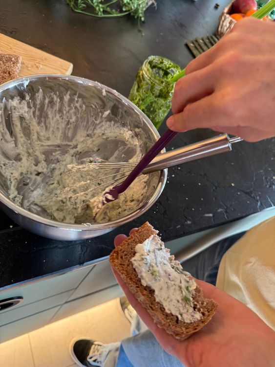 Person bestreicht Kräutercreme auf Stück Roggenbrot in Küche mit Schüssel und Glas mit Kräutern