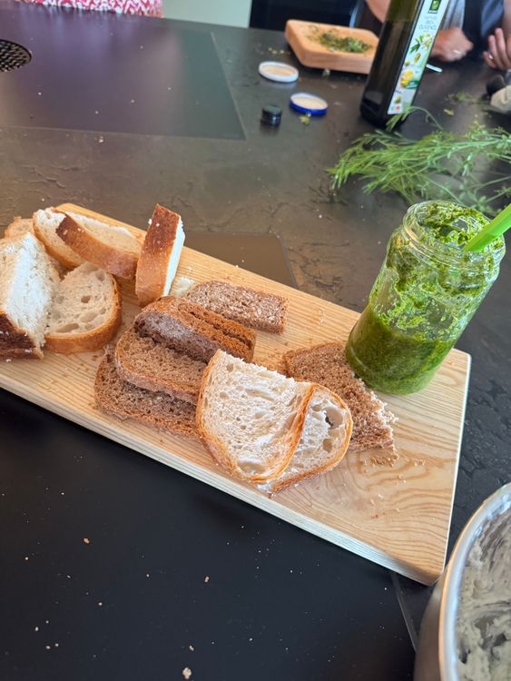 Verschiedene Brotscheiben auf einem Holzbrett mit grünem Pesto im Glas und Kräutern im Hintergrund