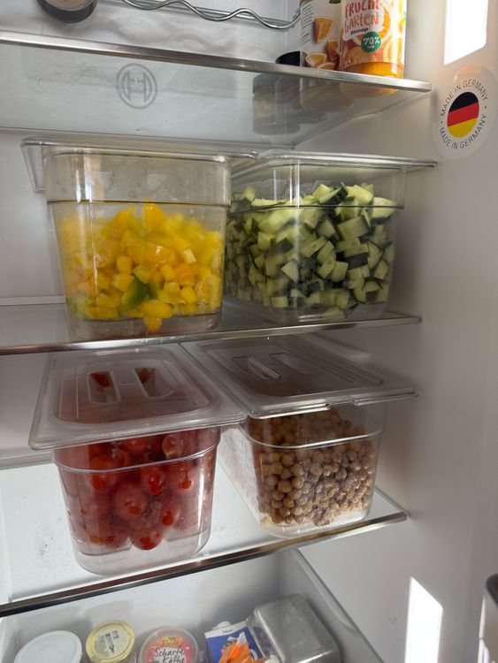 Lebensmittel in durchsichtigen Behältern im Kühlschrank, darunter Paprika, Gurken, Kichererbsen und Tomaten