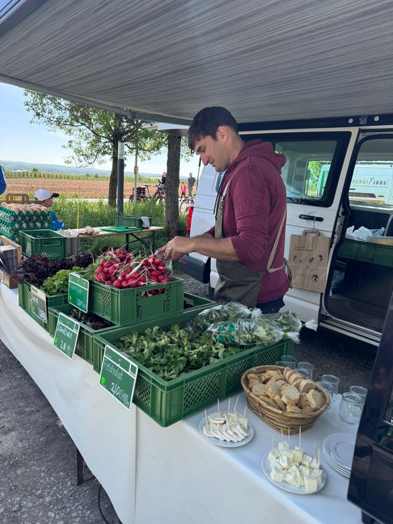 Marktstand mit frischem Gemüse und Verkäufer unter einem Sonnenschutz neben einem weißen Van.