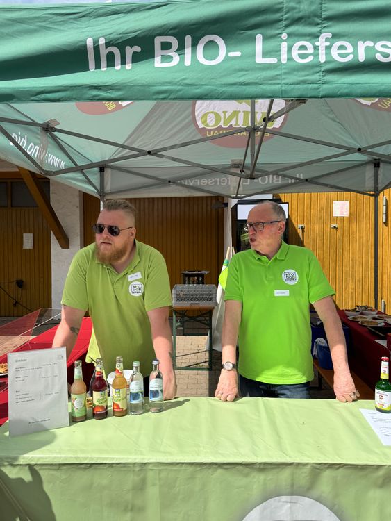 Zwei Männer in grünen Hemden stehen an einem Stand mit Bio-Getränken unter einem Zelt mit dem Aufdruck Bio-Lieferservice