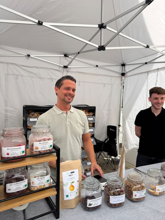 Zwei Männer lächeln an einem Marktstand mit getrockneten Bio-Früchten in Gläsern und Beuteln unter einem weißen Zelt.