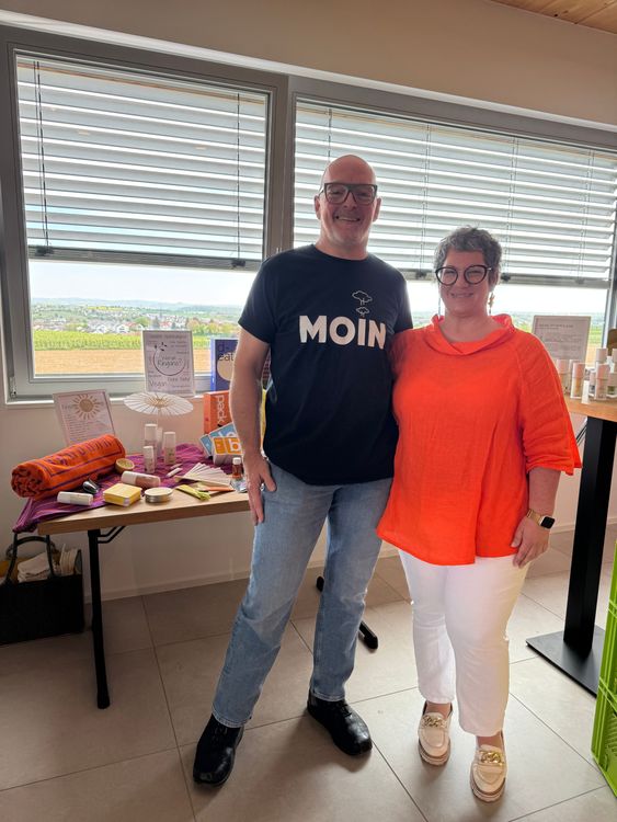 Ein Mann und eine Frau stehen lachend in einem Raum vor einem Tisch mit Kosmetikprodukten und Informationsmaterial.
