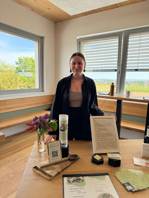 Frau präsentiert veganes Kochbuch und Coaching-Material vor Fenster mit Landschaftsblick in modernem Raum