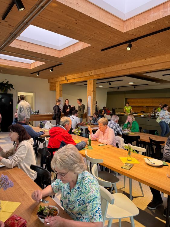Menschen sitzen und essen gemeinsam in einer modernen Cafeteria mit Holzdecke und großen Fenstern.