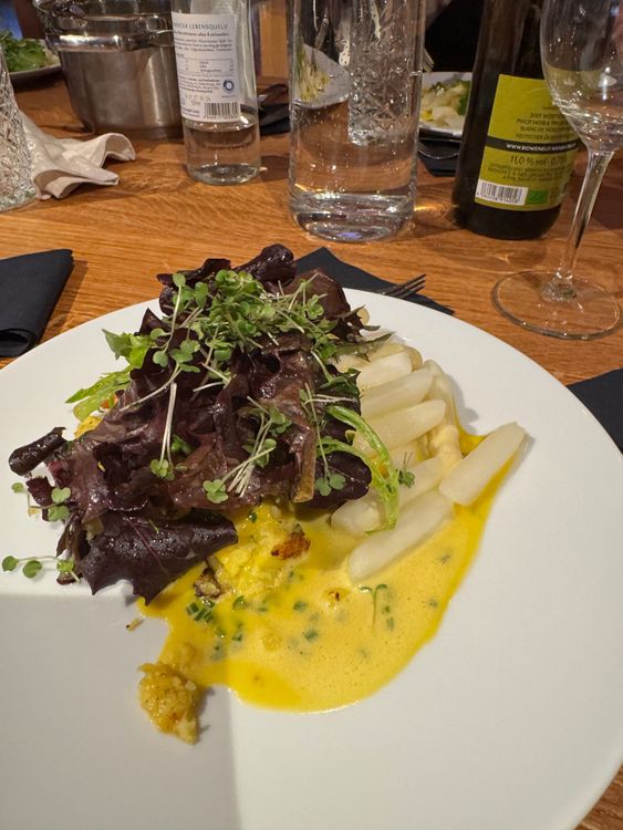 Teller mit weißem Spargel, grünen Salatblättern und gelber Sauce auf Holztisch mit Wasserflaschen und Weinglas