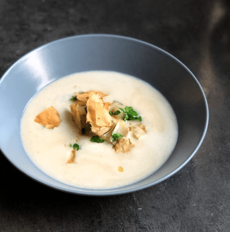 Schüssel mit cremiger weißer Suppe garniert mit Kräutern und knusprigen Chips auf dunklem Untergrund