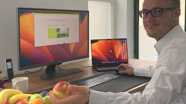 Mann im Büro schaut auf Computer, hält Apfel in der Hand, Obstschale auf dem Tisch.