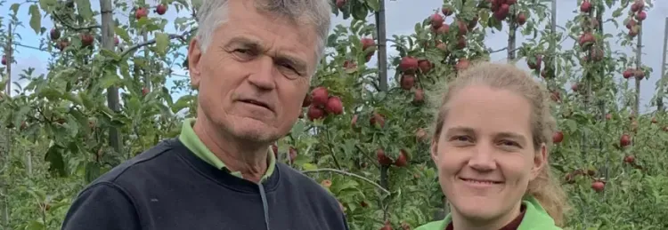 Zwei Personen stehen in einem Apfelgarten mit reifen roten Äpfeln im Hintergrund