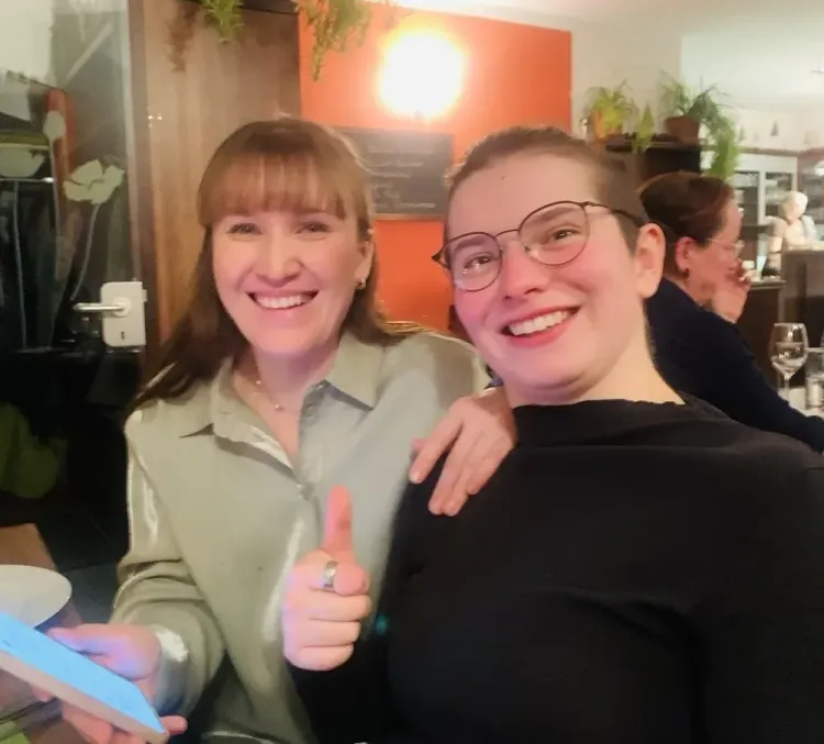 Zwei lächelnde Frauen in einem Restaurant, eine zeigt Daumen hoch, im Hintergrund Gäste und Pflanzen