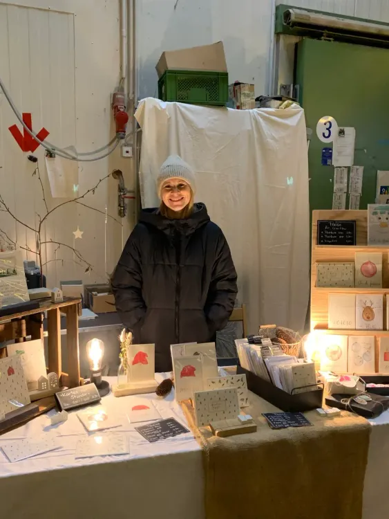 Lächelnde Frau in warmer Jacke und Mütze an Marktstand mit handgemachten Karten und Dekorationen
