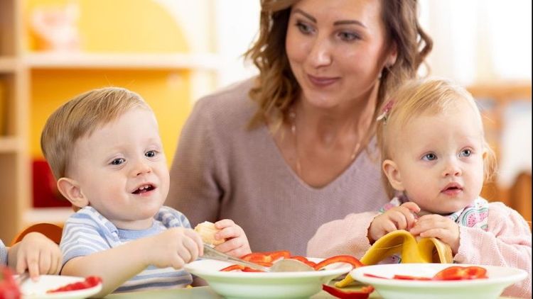 Zwei Kleinkinder essen an einem Tisch mit einer betreuenden Frau im Hintergrund.