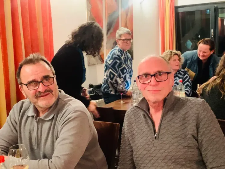 Mehrere Personen sitzen an einem Tisch bei einem geselligen Treffen in einem Raum mit orangen Vorhängen.