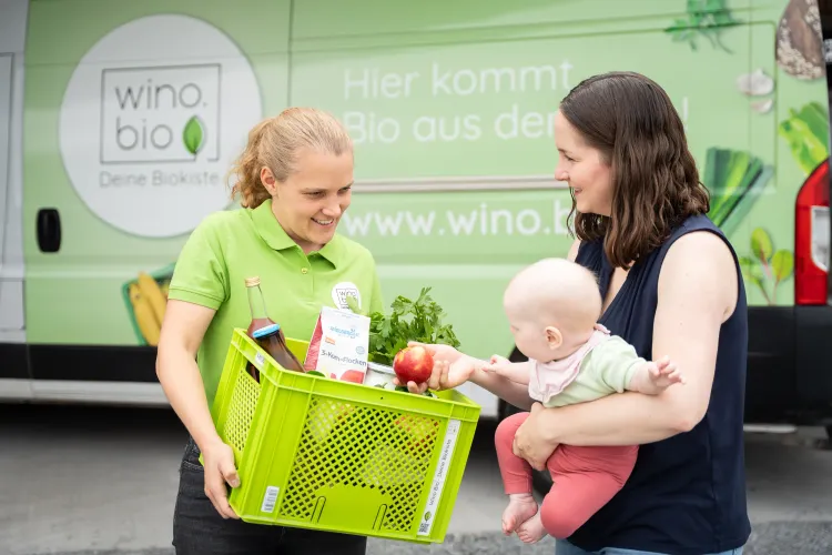 Frau übergibt Bio-Lebensmittelkiste an Mutter mit Baby vor grünem Liefervan von wino.bio