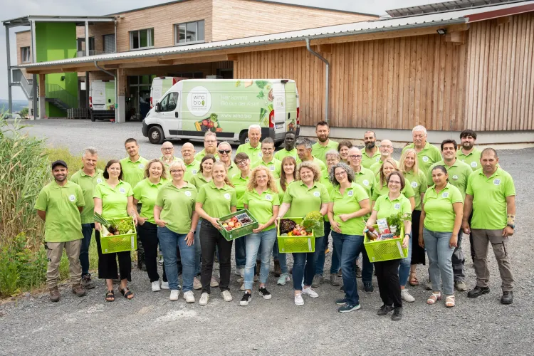Große Gruppe von Menschen in grünen Poloshirts mit Biokisten vor Holzgebäude und Lieferwagen