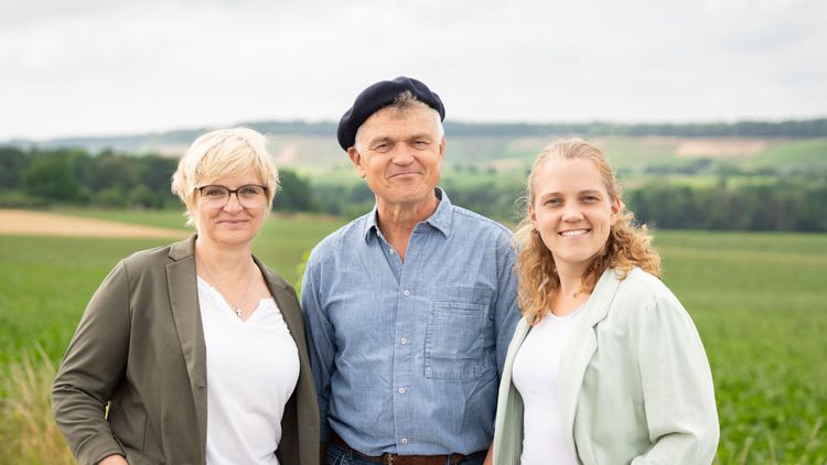 Drei lächelnde Personen stehen auf einem Feldweg vor grünen Feldern.