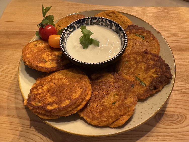 Teller mit goldbraunen Gemüsepuffer und Joghurt-Dip, garniert mit Kirschtomaten und frischem Koriander