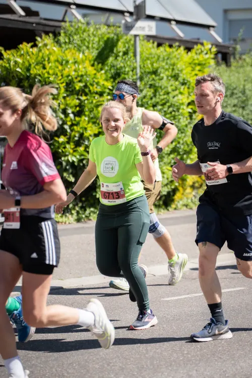 Läuferinnen und Läufer bei einem Straßenlauf im Sonnenschein, eine lächelnde Frau winkt in grüner Sportkleidung.