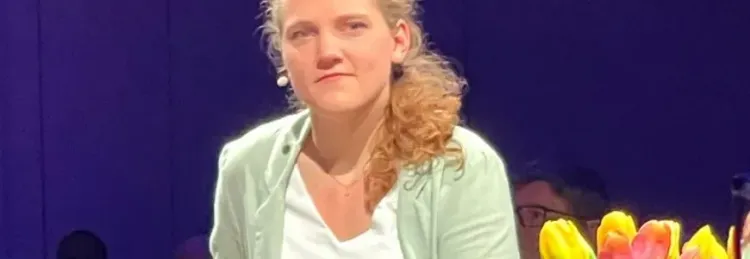 Frau mit lockigem Haar trägt ein hellgrünes Jackett und sitzt vor einem Tulpenstrauß auf dunklem Hintergrund