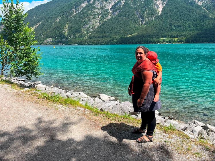 Frau trägt Kind auf dem Rücken am türkisfarbenen See mit Bergen und Wald im Hintergrund