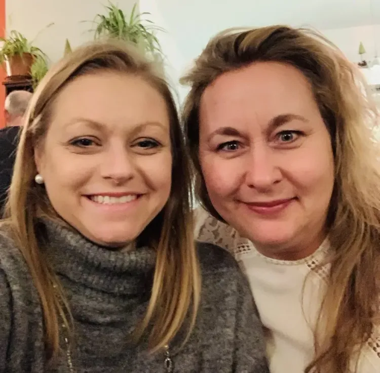Zwei lächelnde Frauen nah beieinander mit Pflanzen im Hintergrund