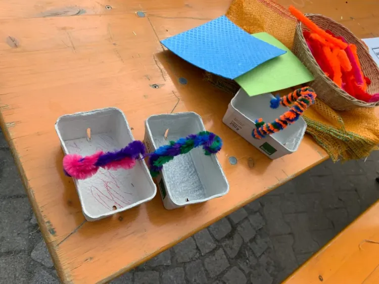 Bunte Pfeifenreiniger in Pappschalen und Bastelmaterial auf einem Holztisch im Freien.
