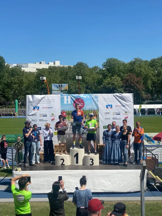 Siegerehrung bei einem Marathon mit drei Läufern auf Podest und Zuschauern bei sonnigem Wetter