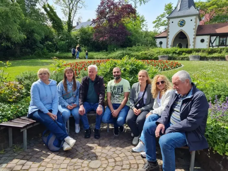 Sieben Personen sitzen auf Bänken im Park vor einem Garten mit Blumen und einer kleinen Kirche im Hintergrund.