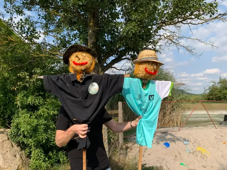 Zwei Vogelscheuchen mit Strohhüten und T-Shirts in einem Garten vor Baum und Sandspielplatz