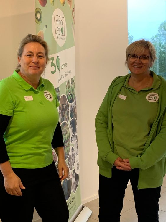 Zwei Frauen in grünen Shirts vor einem Werbebanner von wino.bio feiern 30 Jahre Bio-Kiste
