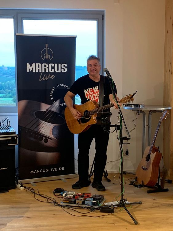 Musiker spielt Gitarre vor Marcus Live Banner bei Wohnzimmerkonzert mit Effektgeräten auf Holzboden