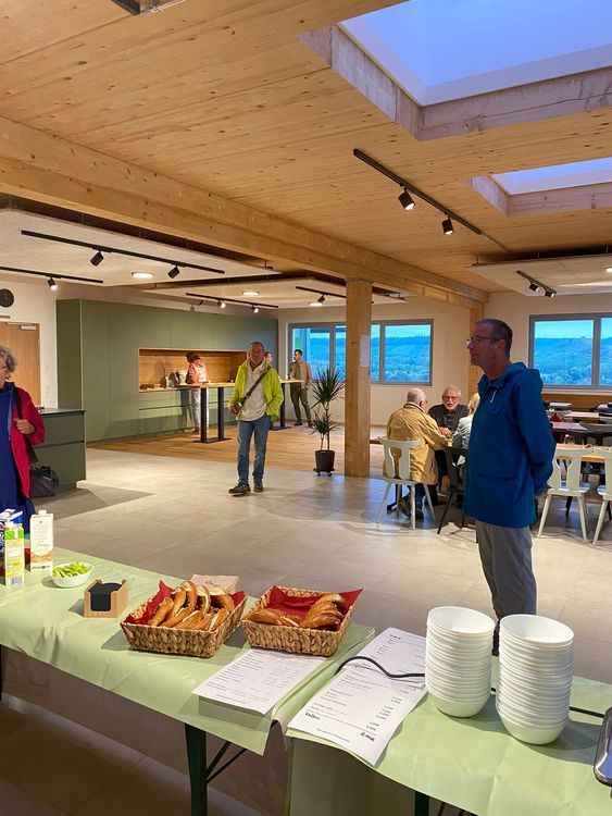 Innenraum mit Holzdach, Tischen, Stühlen und einem Buffet mit Brezeln und Tellern, Menschen im Hintergrund