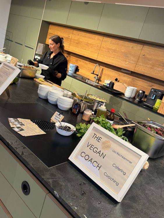 Frau serviert veganes Essen an einer Küche mit The Vegan Coach Schild und Kochutensilien