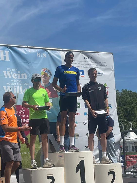 Drei Sportler stehen auf einem Siegerpodest bei einer Siegerehrung im Freien unter blauem Himmel