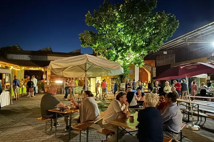 Menschen sitzen abends im Freien an Biertischen unter hell beleuchtetem Baum und Sonnenschirmen
