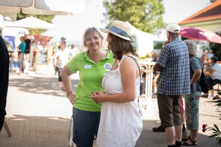 Zwei Frauen sprechen auf einem sonnigen Außenmarkt, eine trägt grünes Polo, die andere weißen Sommerkleid und Hut