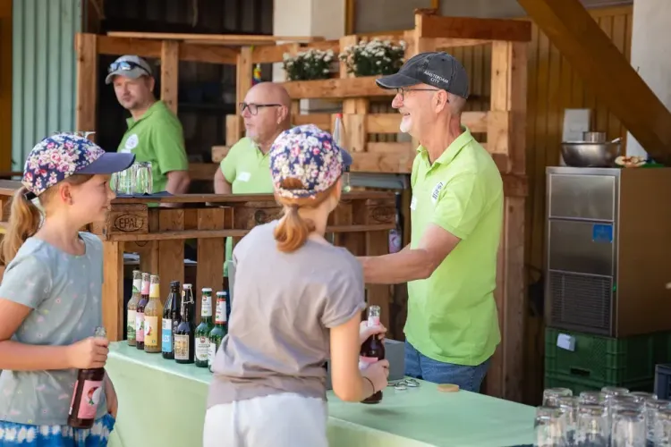 Kinder kaufen Getränke an einem Stand, betreut von Männern in grünen Poloshirts mit Kappen.