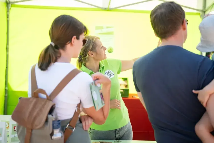 Frau im grünen Poloshirt erklärt zwei Erwachsenen und Kind etwas auf einem Marktstand