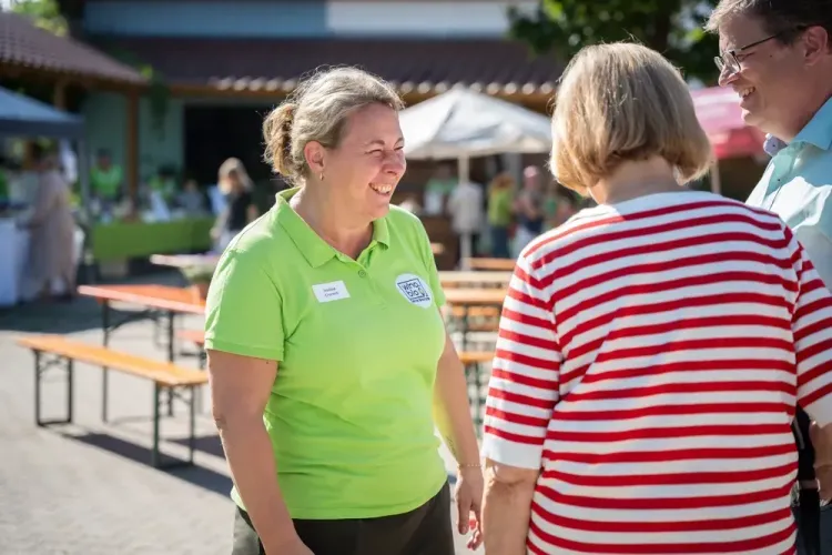 Frau im grünen Polo lacht im Gespräch mit zwei Personen bei sonnigem Outdoor-Event mit Bänken und Ständen