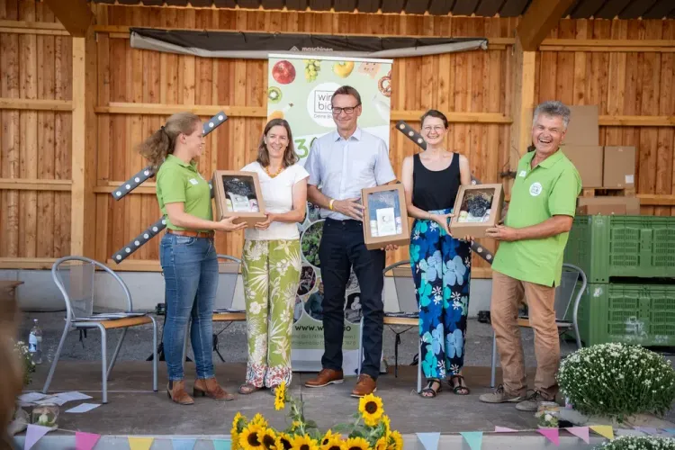 Fünf Personen stehen auf einer Bühne und zeigen Geschenkboxen, Sommerveranstaltung mit Sonnenblumen und Holzwand im Hintergrund