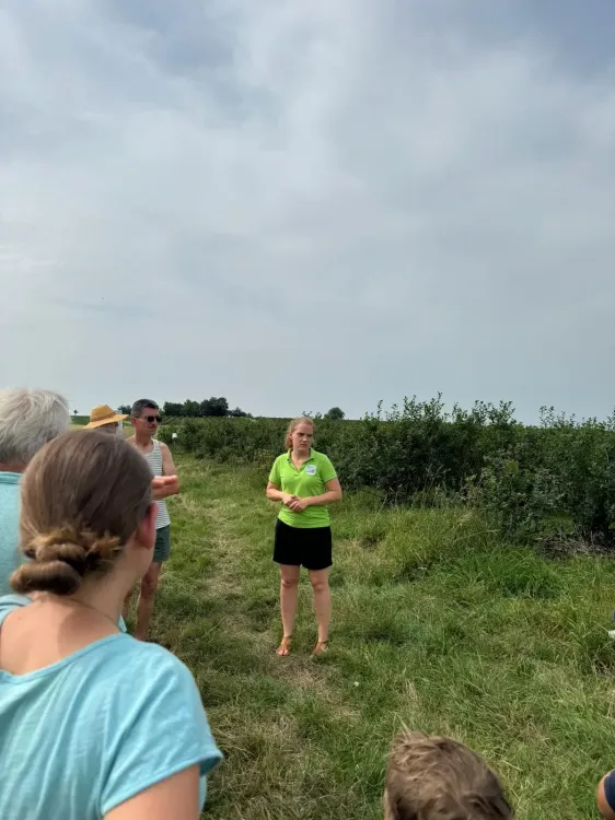 Frau in grünem Hemd erklärt Gruppe von Menschen auf einem Feld mit Büschen unter bewölktem Himmel