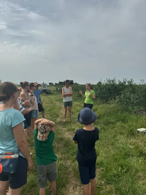 Eine Gruppe Menschen, darunter Kinder, hören einer Frau im Freien auf einem Feld zu.