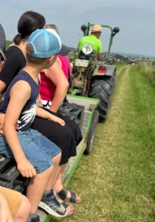 Mehrere Personen sitzen auf einem Anhänger hinter einem Traktor auf einem Feldweg.