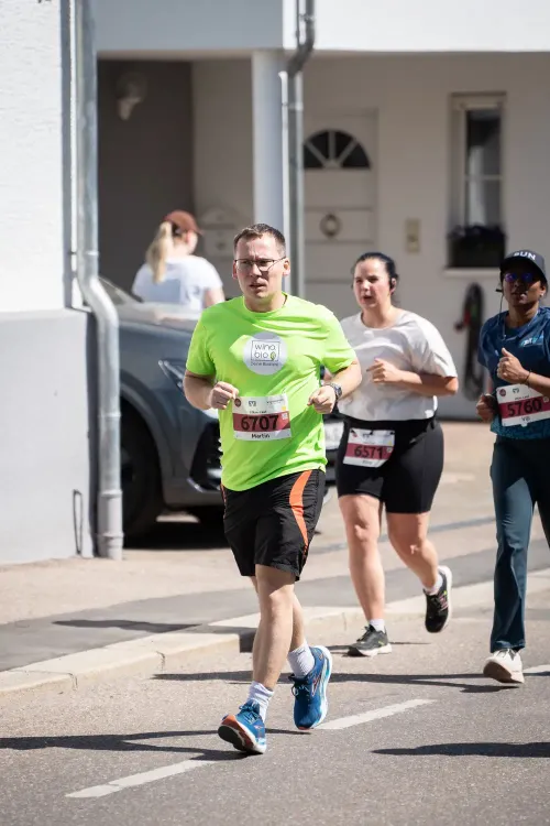 Mehrere Läufer, darunter ein Mann in grünem Shirt mit Startnummer, bei einem Straßenlauf an einem sonnigen Tag