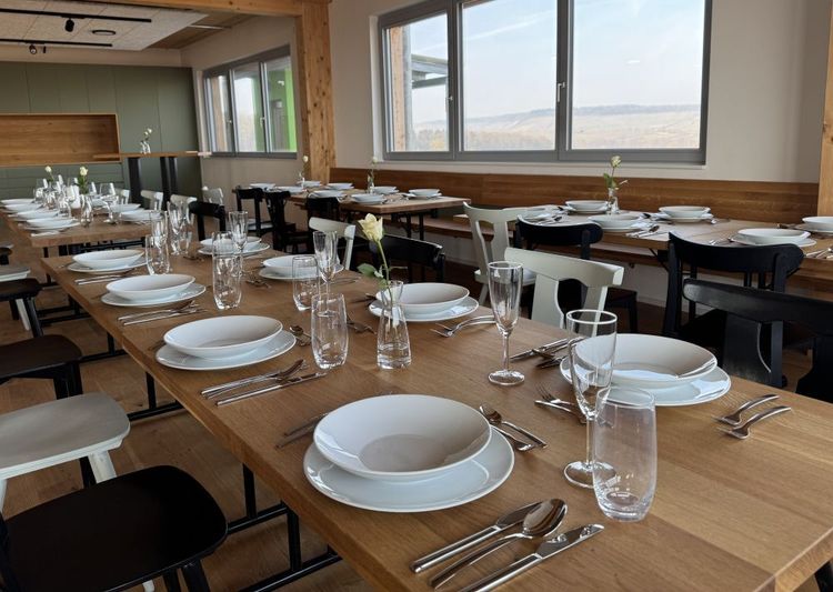 Gedeckte Holztische in einem Restaurant mit weißen Tellern, Gläsern und Blumenvasen neben großen Fenstern.