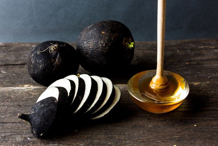 Schwarzer Rettich, ganz und in Scheiben geschnitten, neben Honig im Glas mit Honiglöffel auf Holzoberfläche.